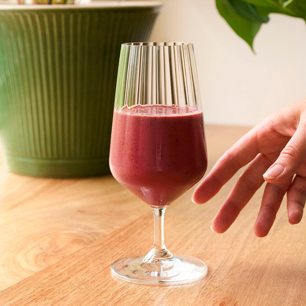 A dark pink smoothie in a fluted glass. A hand is reaching in to pick up the smoothie.